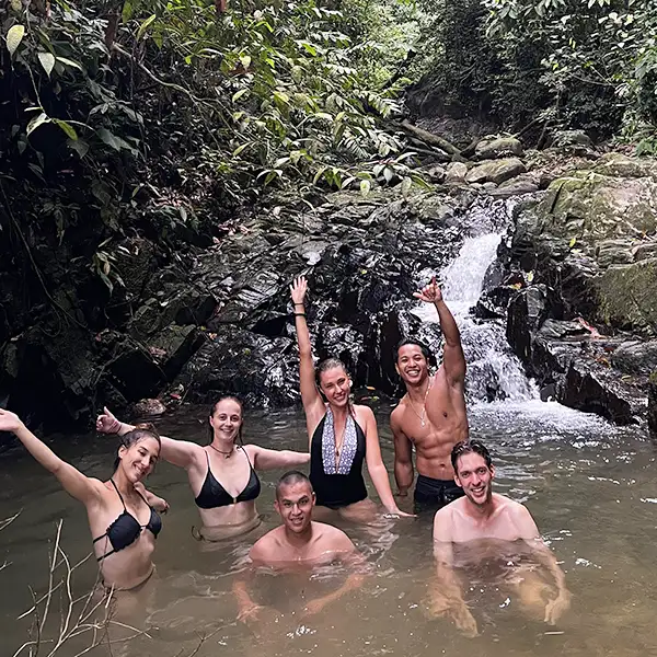 Joyful trekkers in swimwear raising their arms in celebration in a natural jungle pool with a small waterfall in the background.