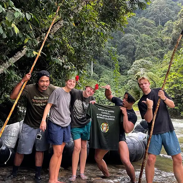 joyful trekking guides and tourists posing with humorous gestures beside a river in a dense forest, one holding a sign with a playful warning about following the guides or getting lost in 'monkey business'.