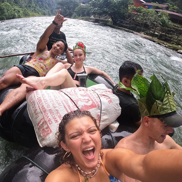 Exuberant rafters taking a selfie on a river raft, with one playfully pointing upwards and another with leaves on their head, embodying the fun of the adventure.
