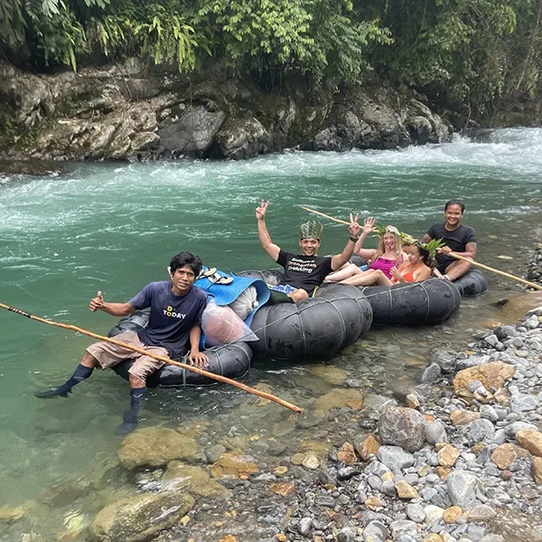 Adventurers on a makeshift raft waving and smiling as they navigate a clear mountain river surrounded by lush greenery.