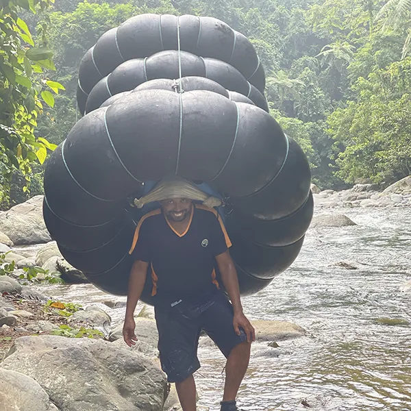 A guide carrying a large stack of black river tubes on his back by a rocky stream, showcasing the behind-the-scenes effort in adventure trekking.