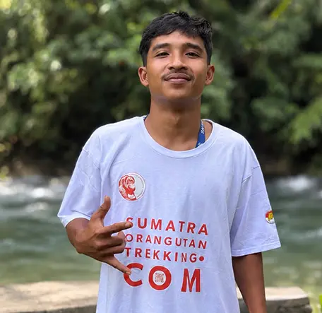 Adit standing beside the river and making a playful hand gesture in a Sumatra Orangutan Trekking T-shirt.