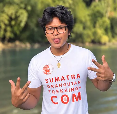 Azizi wearing glasses and posing with both hands raised beside the river.
