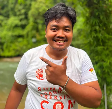 Indra smiling and pointing to the side beside the river, with green foliage behind him.