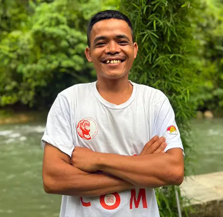 Malem smiling with his arms folded beside the river, with bamboo behind him.