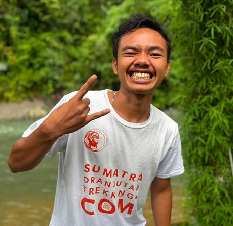 Rahmat grinning and making a playful hand gesture beside the river.