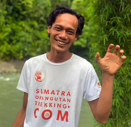 Zaky smiling and waving beside the river, with bamboo behind him.