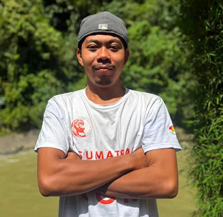 Austin wearing a backwards cap and standing with his arms folded beside the river.
