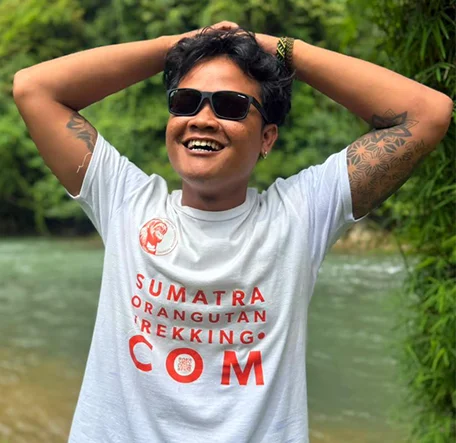 Enu wearing sunglasses and smiling with both hands resting on his head beside the river.
