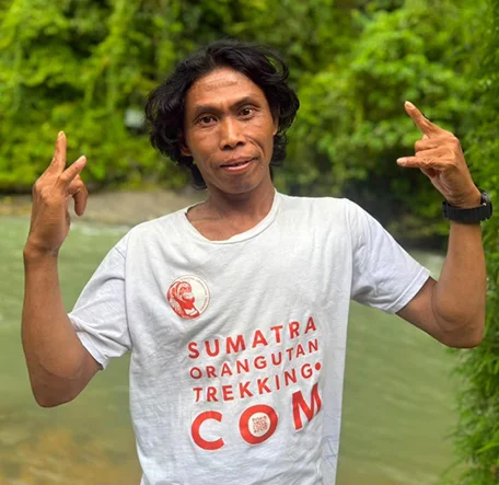 Memet posing with both hands raised beside the river in a Sumatra Orangutan Trekking T-shirt.