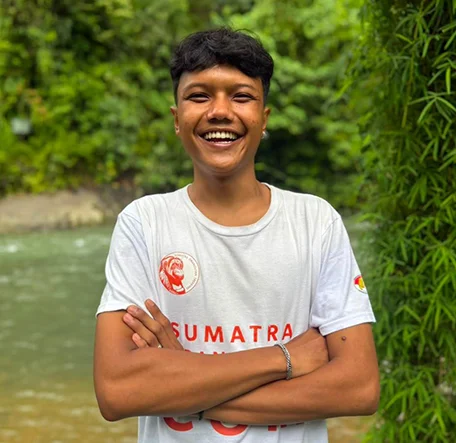 Rizky smiling with his arms folded beside the river in a Sumatra Orangutan Trekking T-shirt.