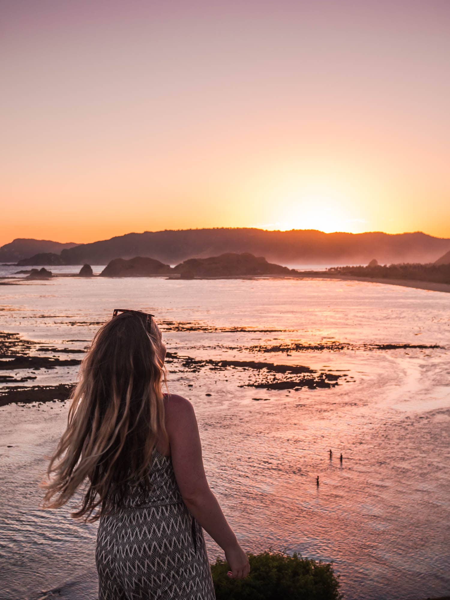 Girl with long hair flowing in the wind, wearing a black and white jumpsuit, is standing on the edge of Bukit Merese at sunset, one of the top things to do in Lombok.