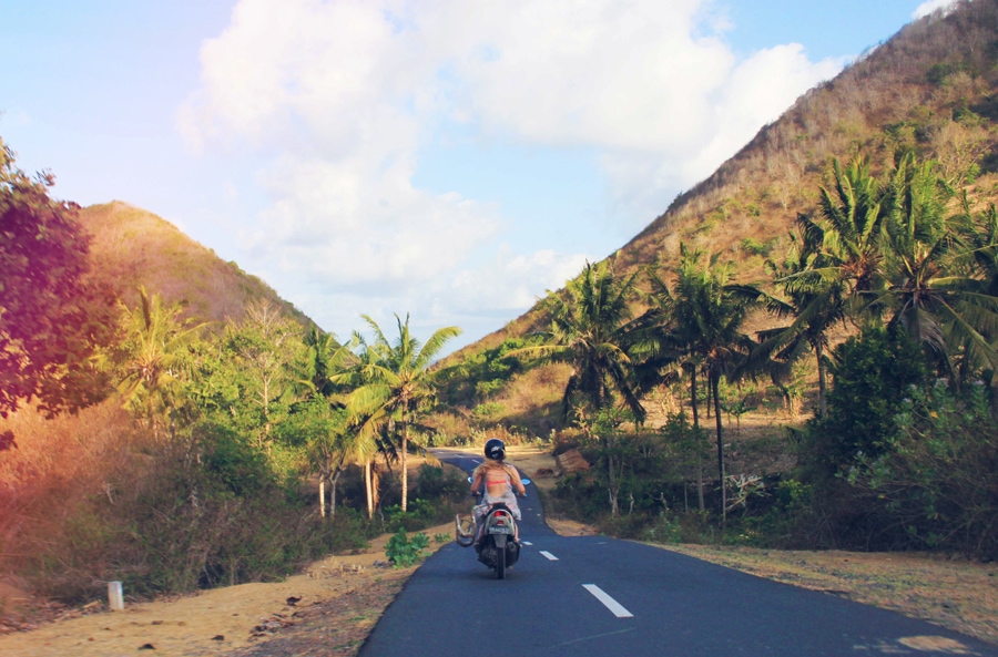 Girls wearing a grey dress and black helmet driving on a narrow road surrounded by palm trees, one of the top things to do in Lombok.