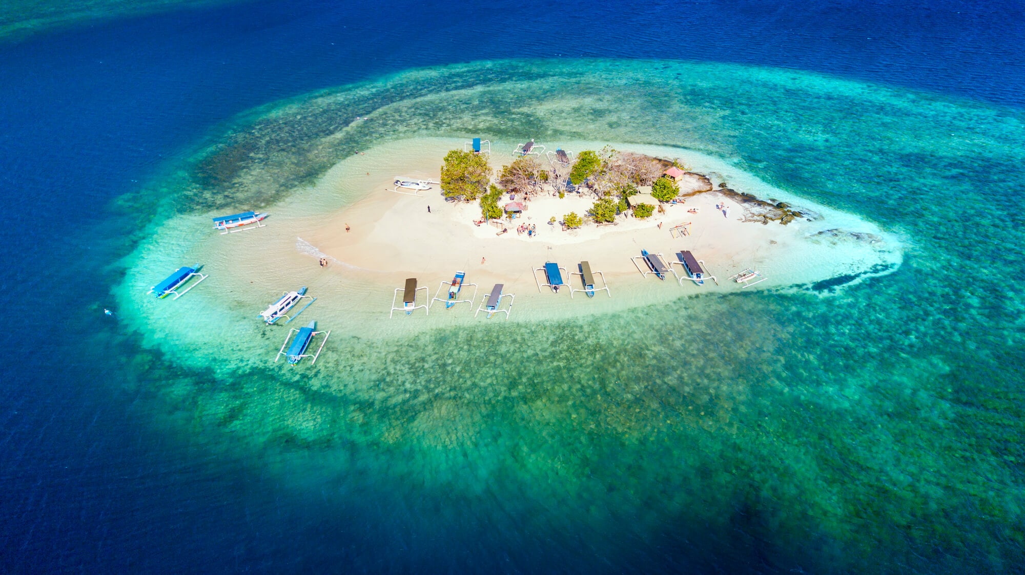 A sandbar surrounded by coral reef and dark blue water with a few traditional Indonesian boats and trees, seen from above.