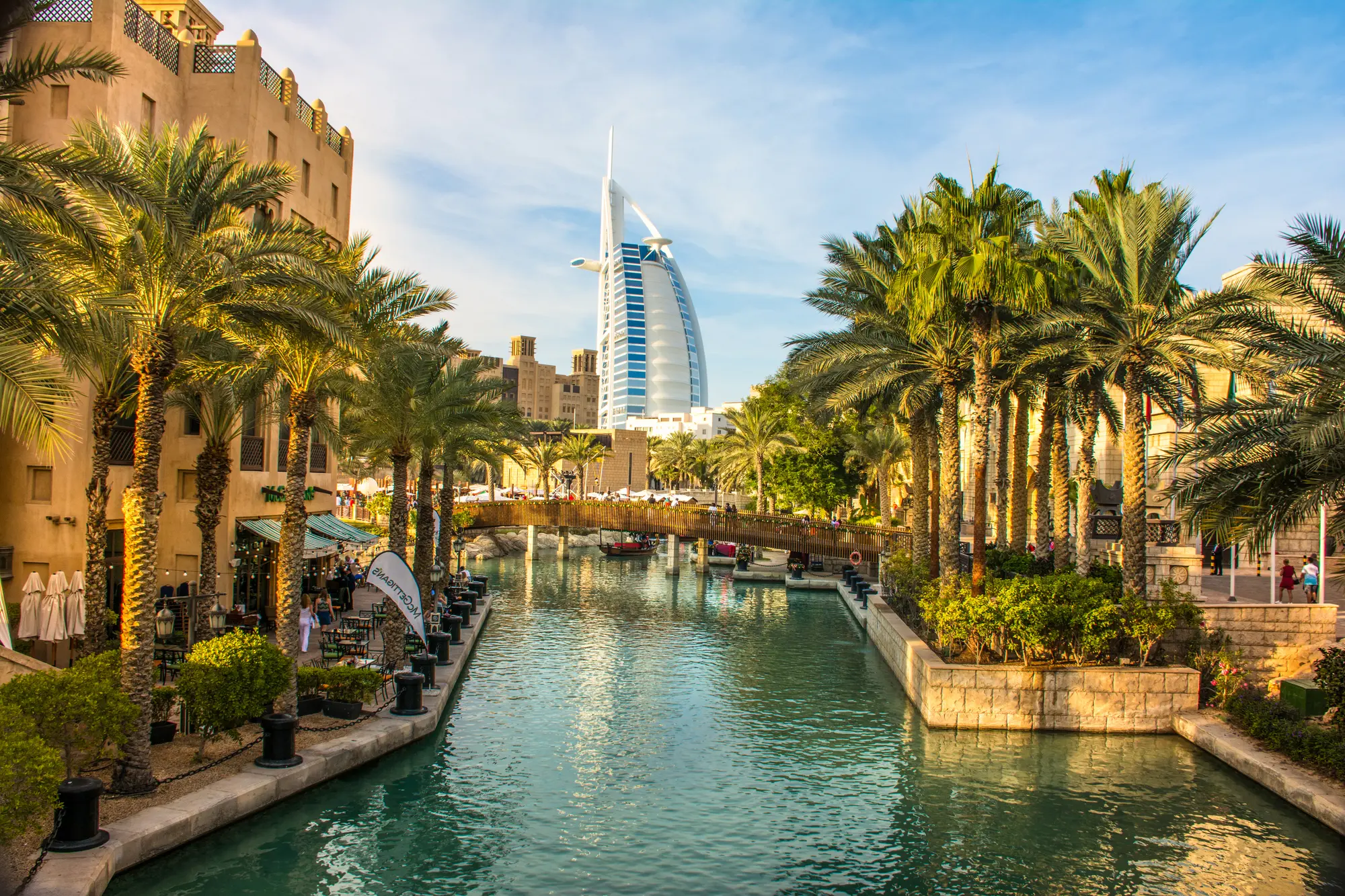 Green canal lined by palm trees and brown traditional Arab houses with Burj Al Arab in the background, must during your 2 days in Dubai.