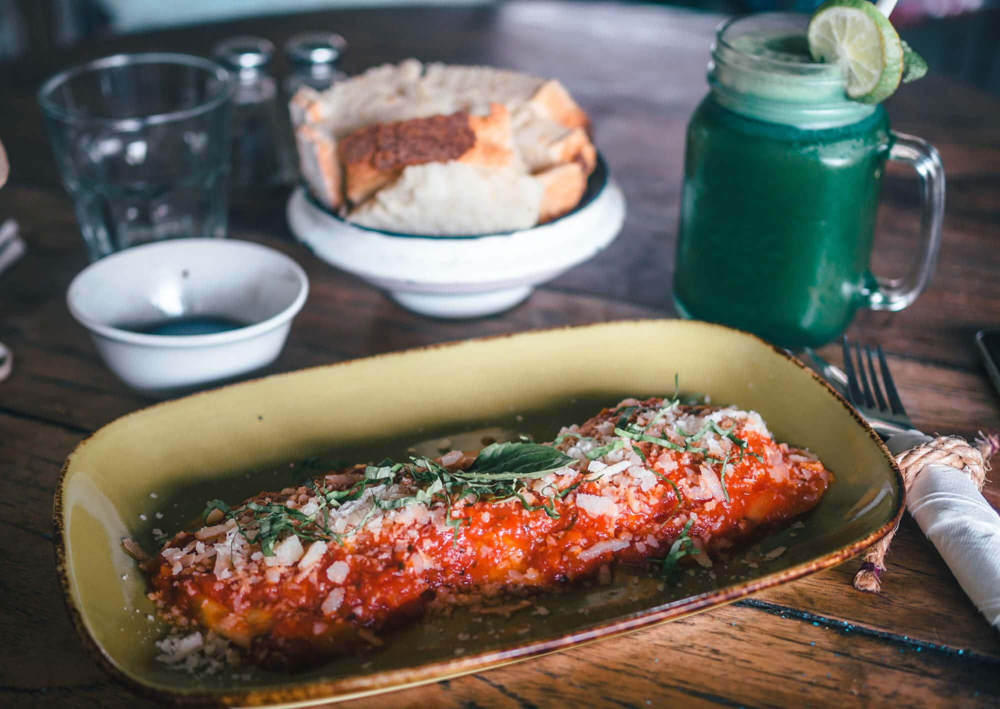 Close up of eggplant parmigiana on a light yellow rectangular plate with a bowl of bread and a green juice in the background at La Baracca, one of the best restaurants in Echo Beach Canggu.