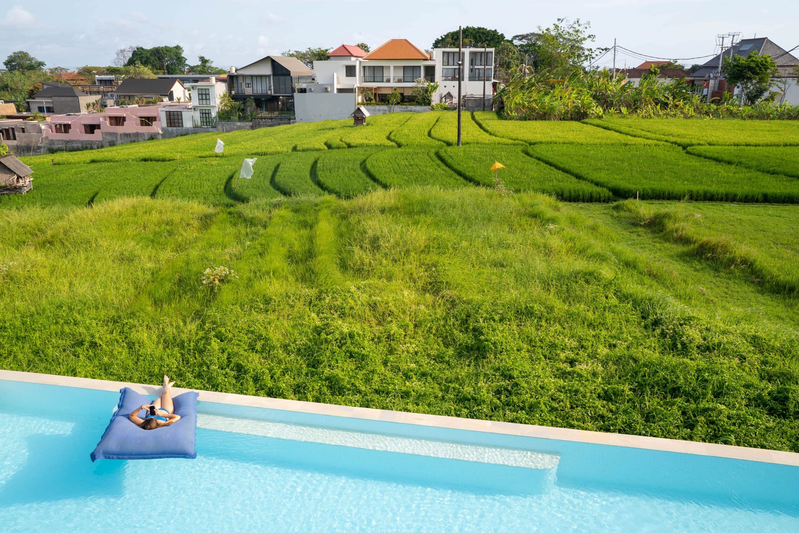 Woman waering a blue bikini lying on a purple floatie on the edge of a pool overlooking lush rice fields at Valle in the Echo Beach area of Canggu