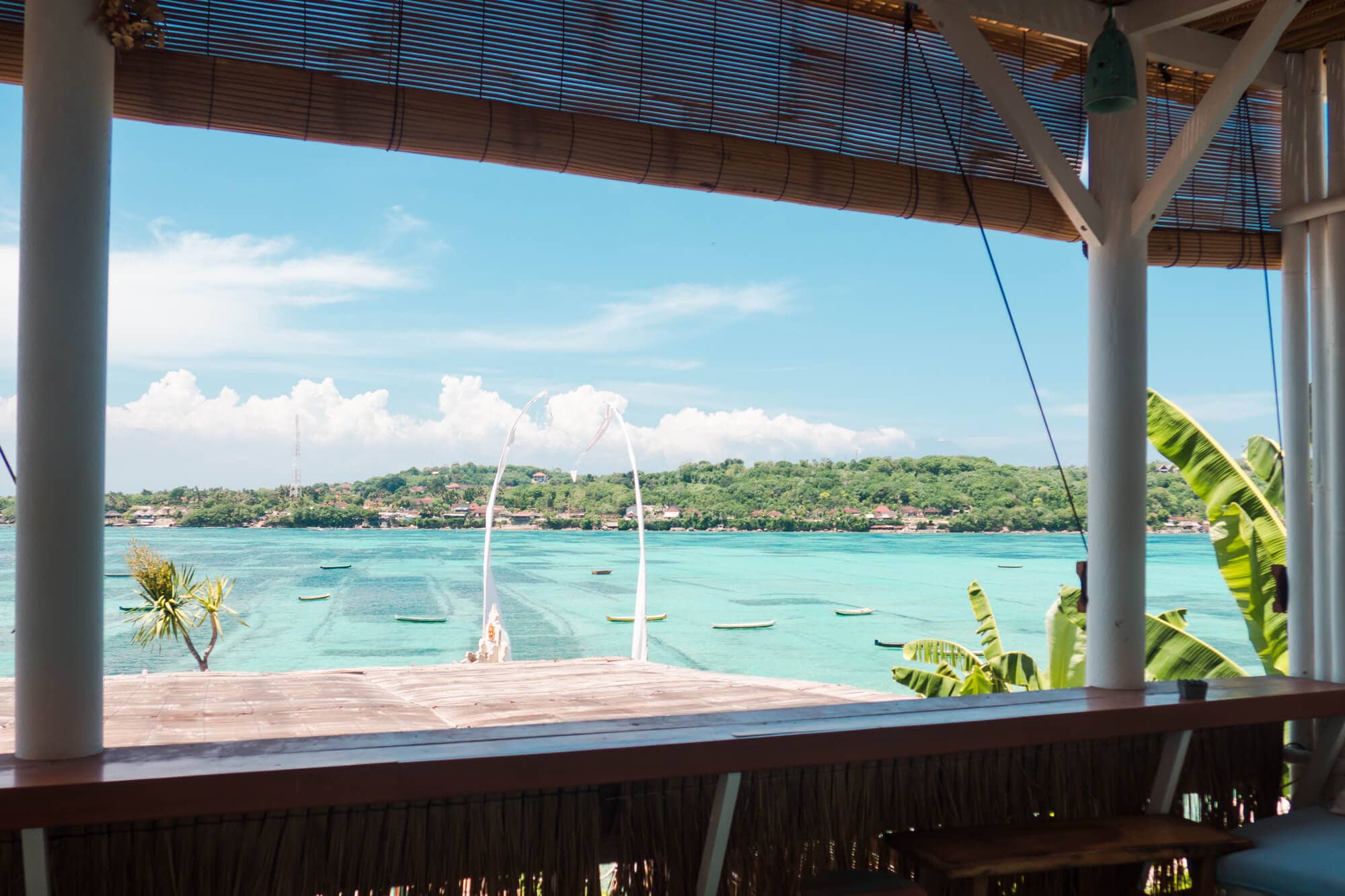 View through a window in the restaurants at Le Pirate Beach Club Nusa Ceningan, overlooking a shallow turquoise lagoon and Lembongan on the other side.