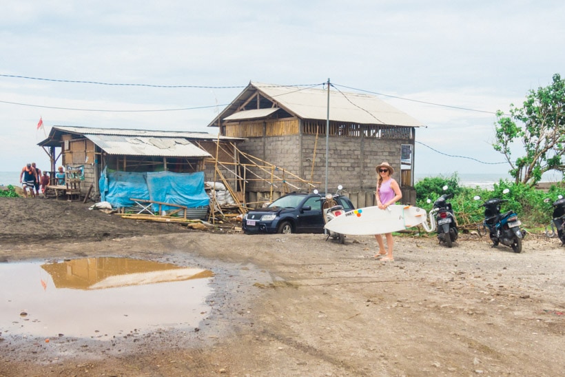 Canggu Echo Beach girl wearing a purple dress and beige hat holding a white surfboard in front of a warung.