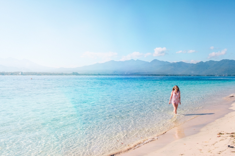 Woman wearing a pink jumpsuit walking along the water's endge on a beach in Gili Air, complete Gili Islands comparison.