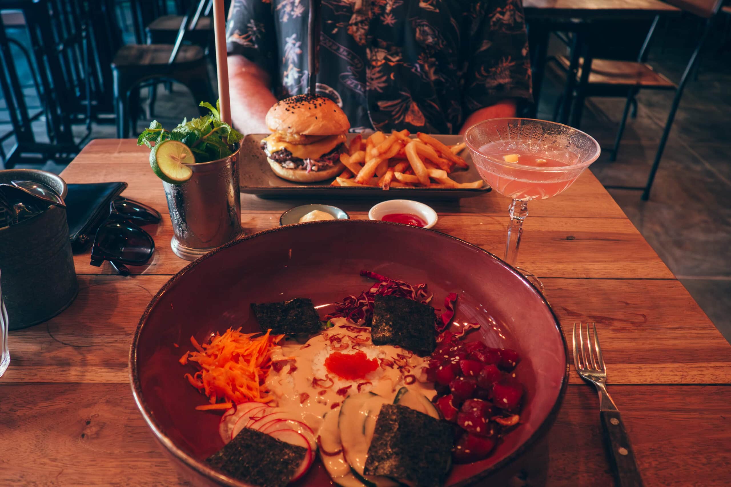 Poké in a red bowl on a wooden table with a pink cocktail and burger with fries in the background at KRNK, one of the best restaurants in Kuta Lombok.