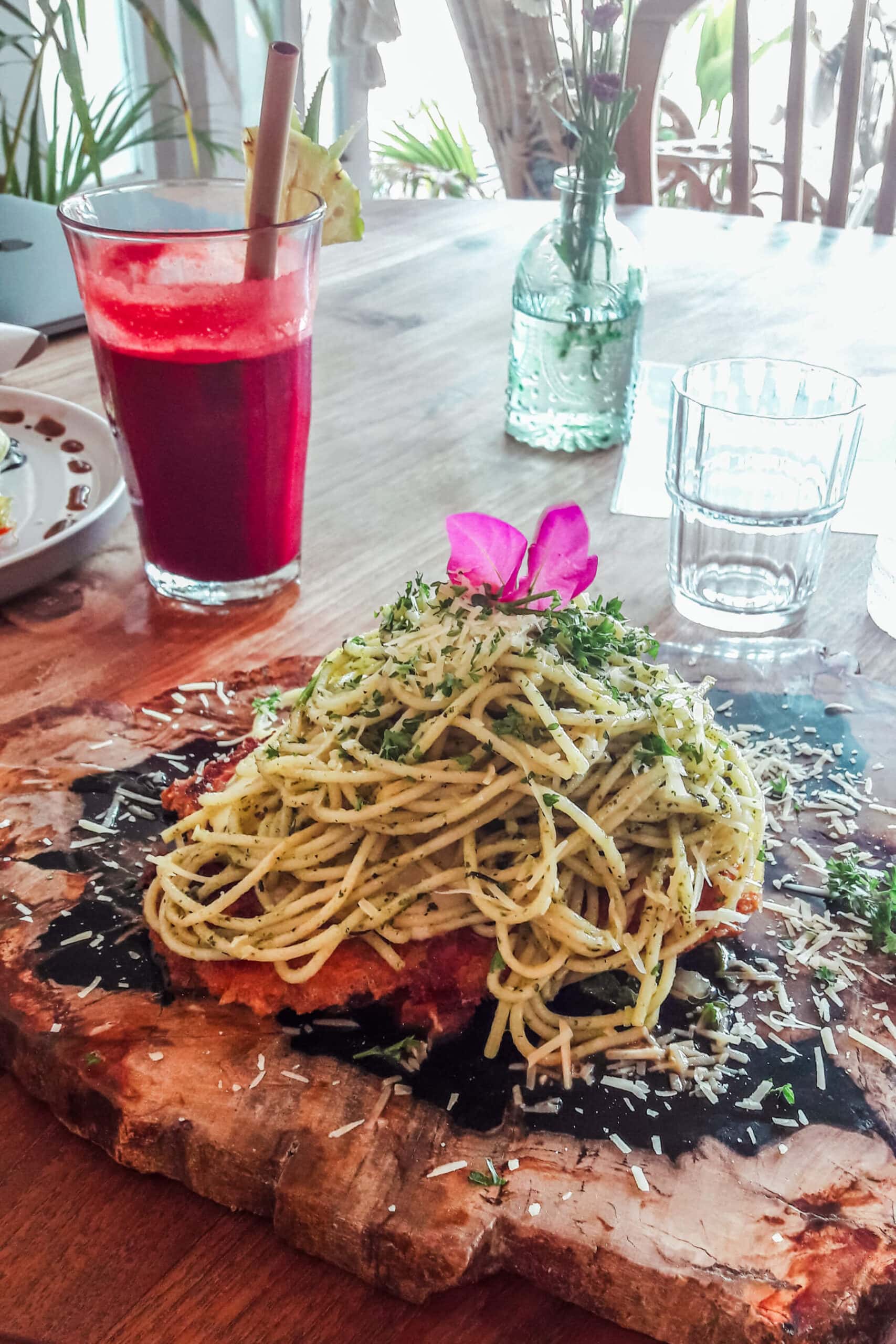 Chicken Parma with spaghetti on a wooden board with a purple flower and a red fruit juice in the background, at Kenza one of the best restaurants in Kuta Lombok.