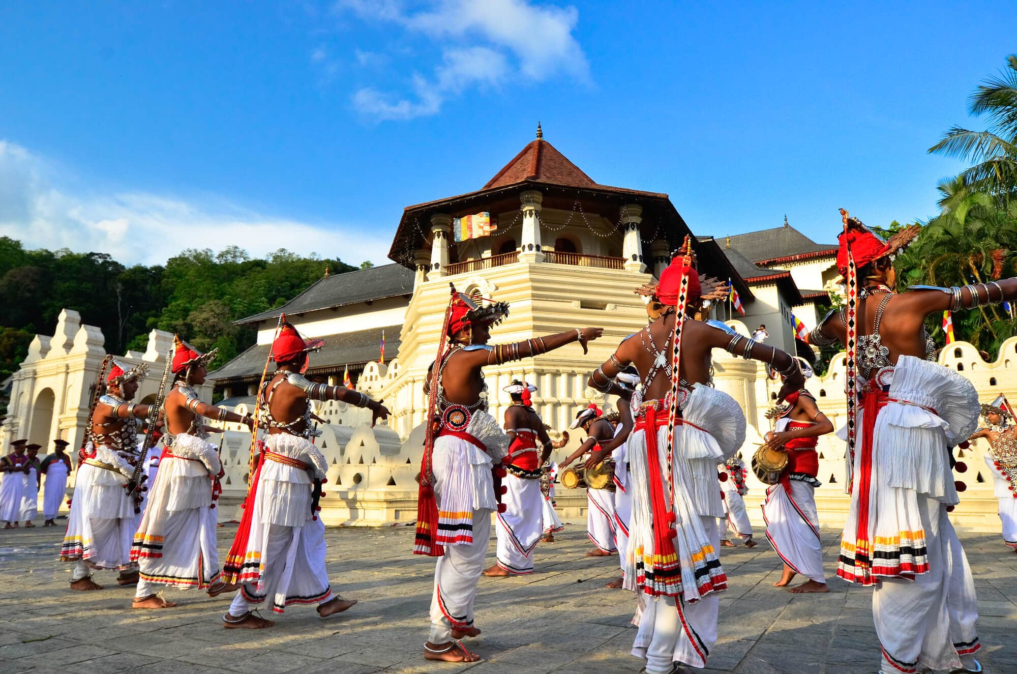 Men wearing white costumes with red hats and jewelry dancing traditional Kandyan Dance outside Temple of the Tooth, one of the best things to see in Kandy.