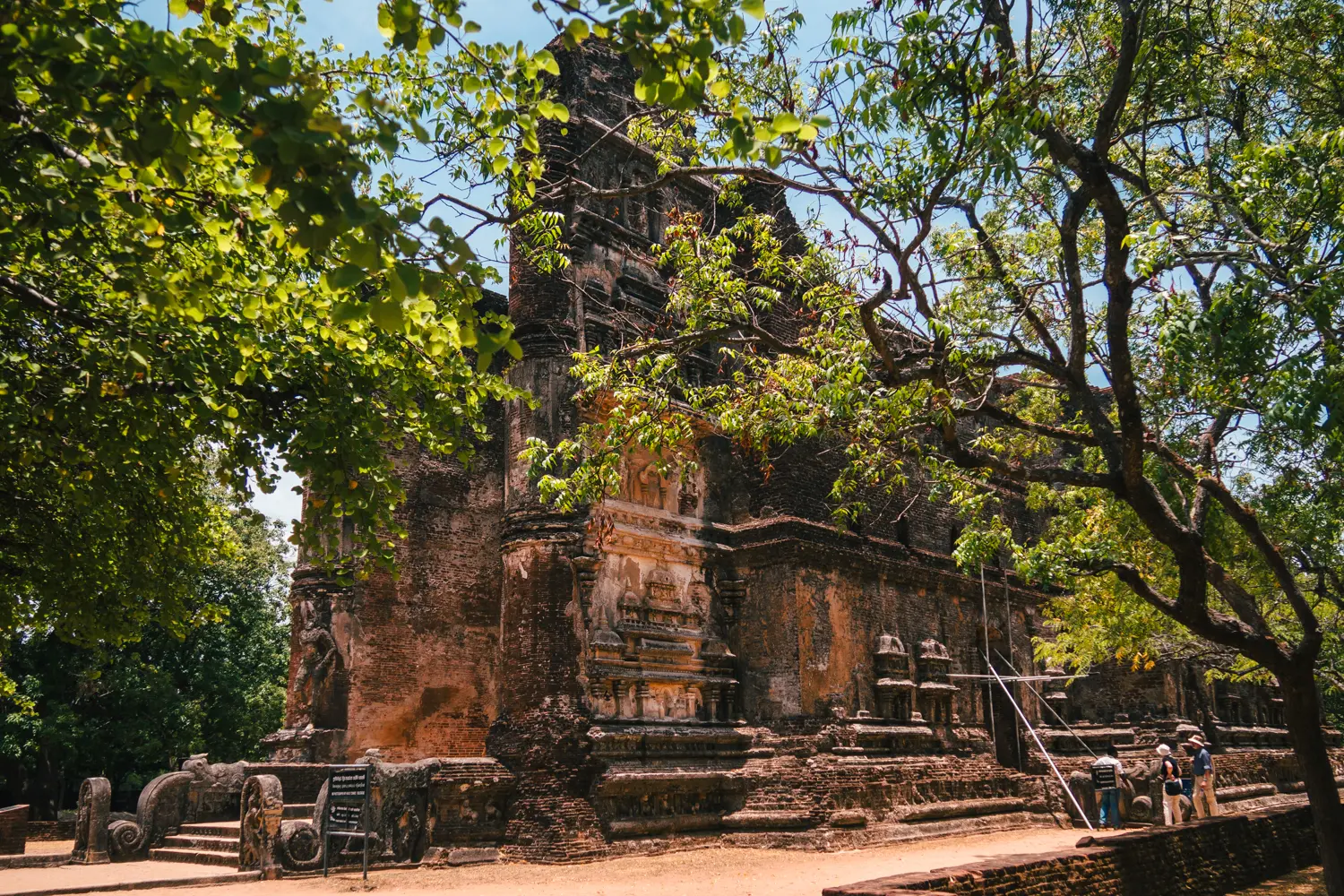 The ornate brown brick ruins of Lankatilaka seen through green trees with three people looking up at it in Polonnaruwa.