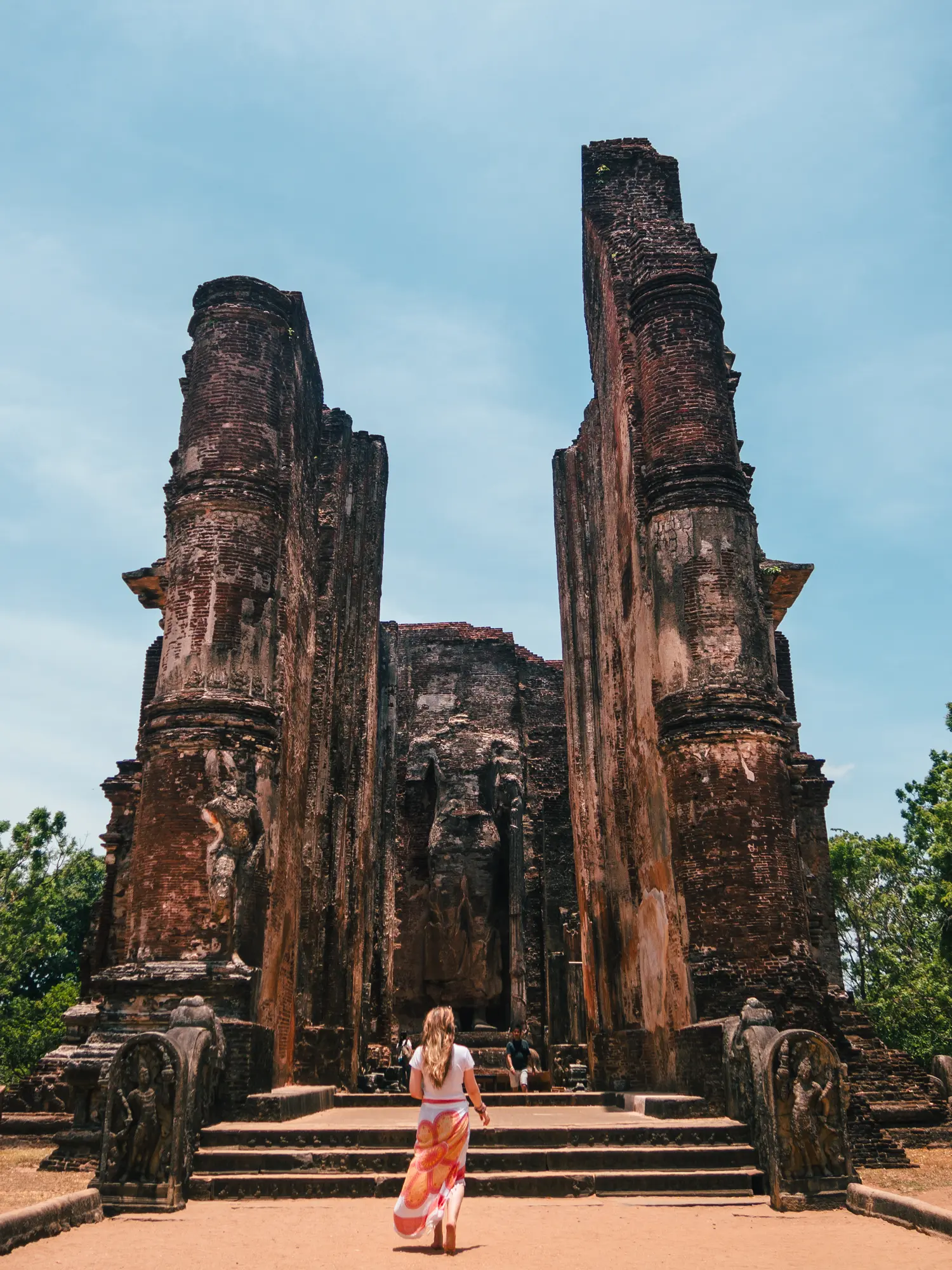 Girl with long hair, wearing a white, orange and yellow sarong, walking towards two tall brick walls with a Buddha statue in the middle, at Lankatilaka in Polonnaruwa.