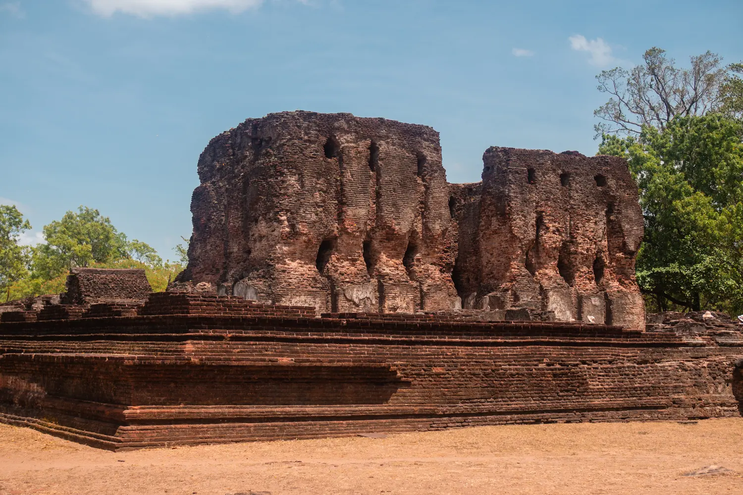 Two brick walls on a square brick foundation surrounded by green trees set against a blue sky, in the ancient city of Polonnaruwa.