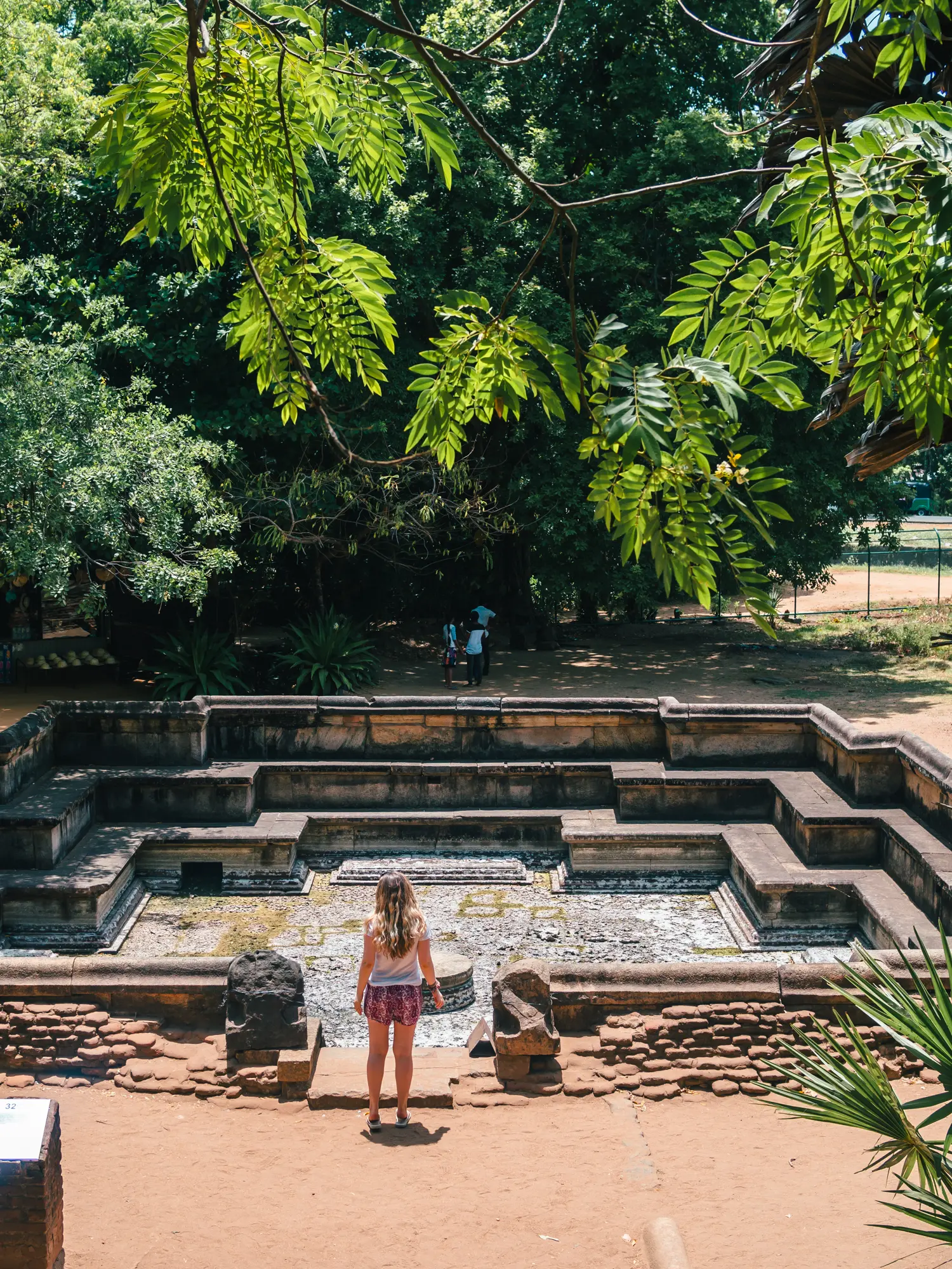 Girl with long hair, wearing a white t-shirt and pink shorts standing in front of the empty stone pool under a green canopy in Polonnaruwa.