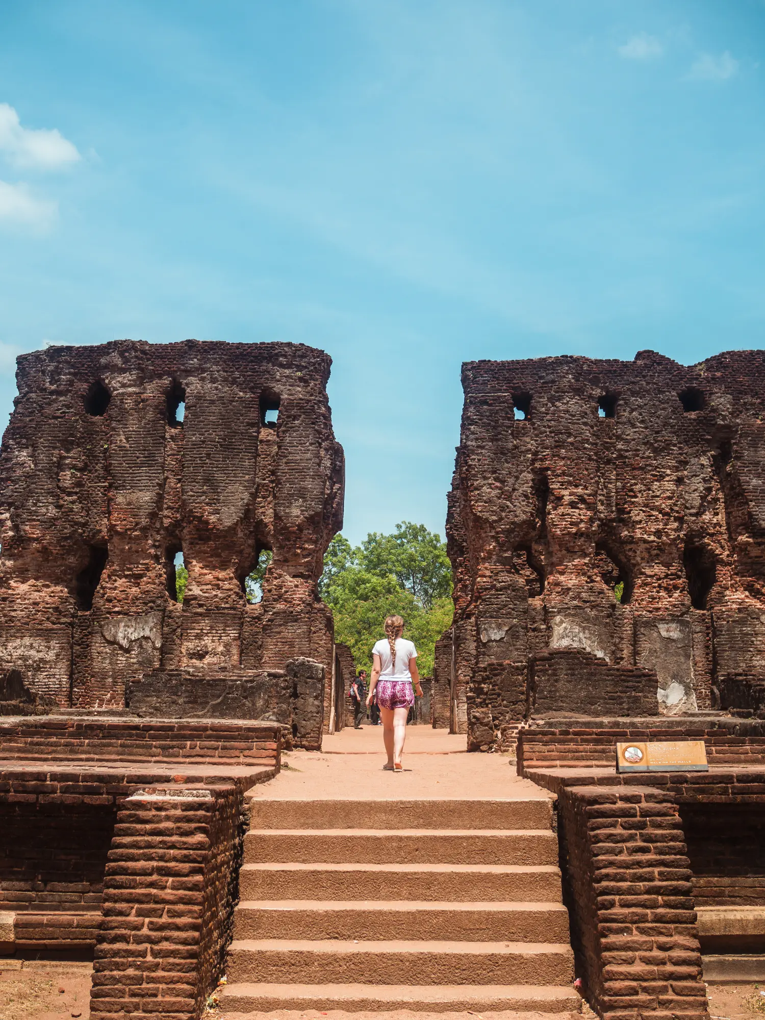Girl in a white t-shirt and pink shorts walking between two brick walls on a sunny day at the Royal Palace ruins in Polonnaruwa.