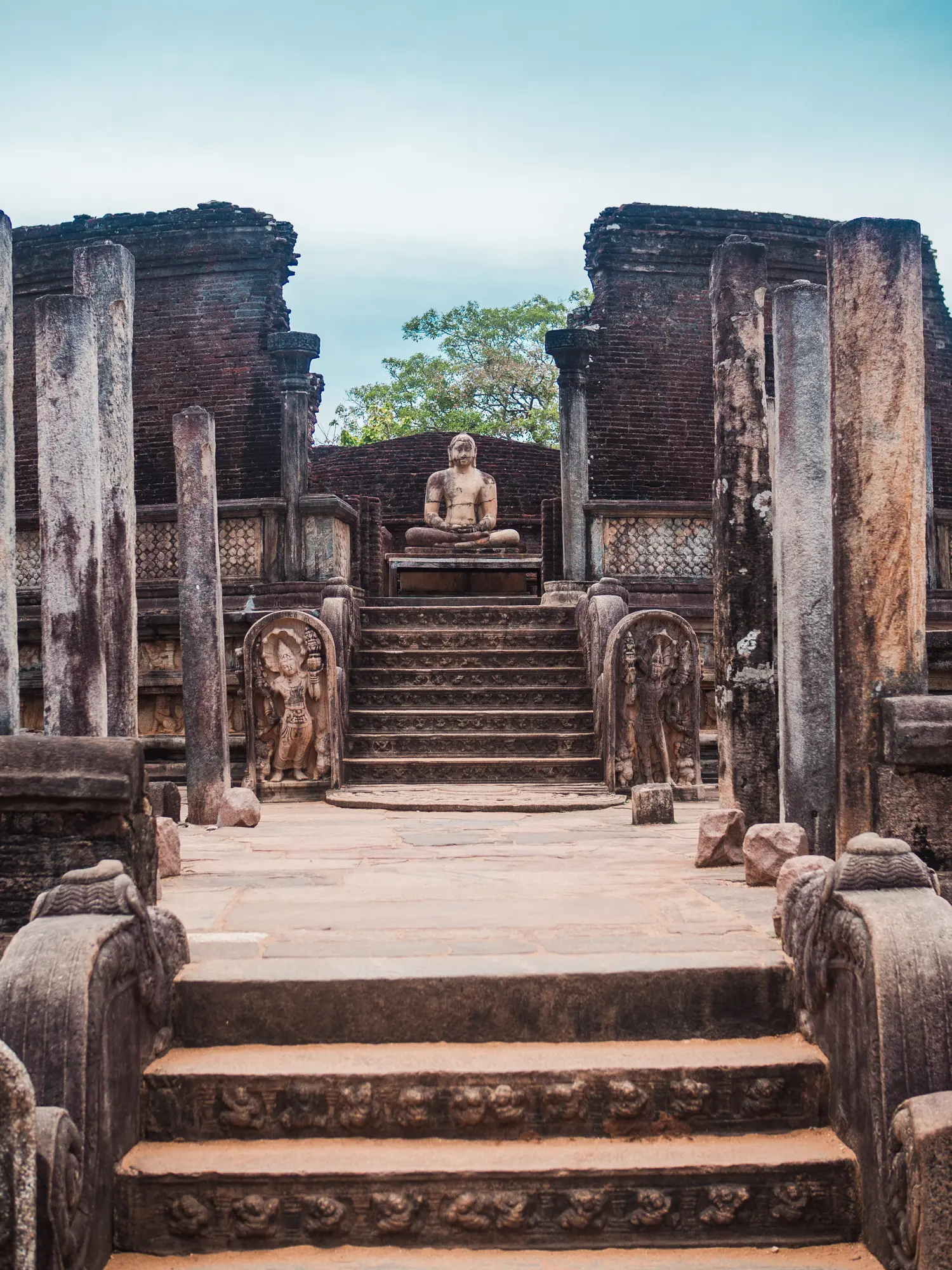 Large stone Buddha sitting at the top of stairs, surrounded by a circular brick wall, in the Sacred Quadrangle in Polonnaruwa.