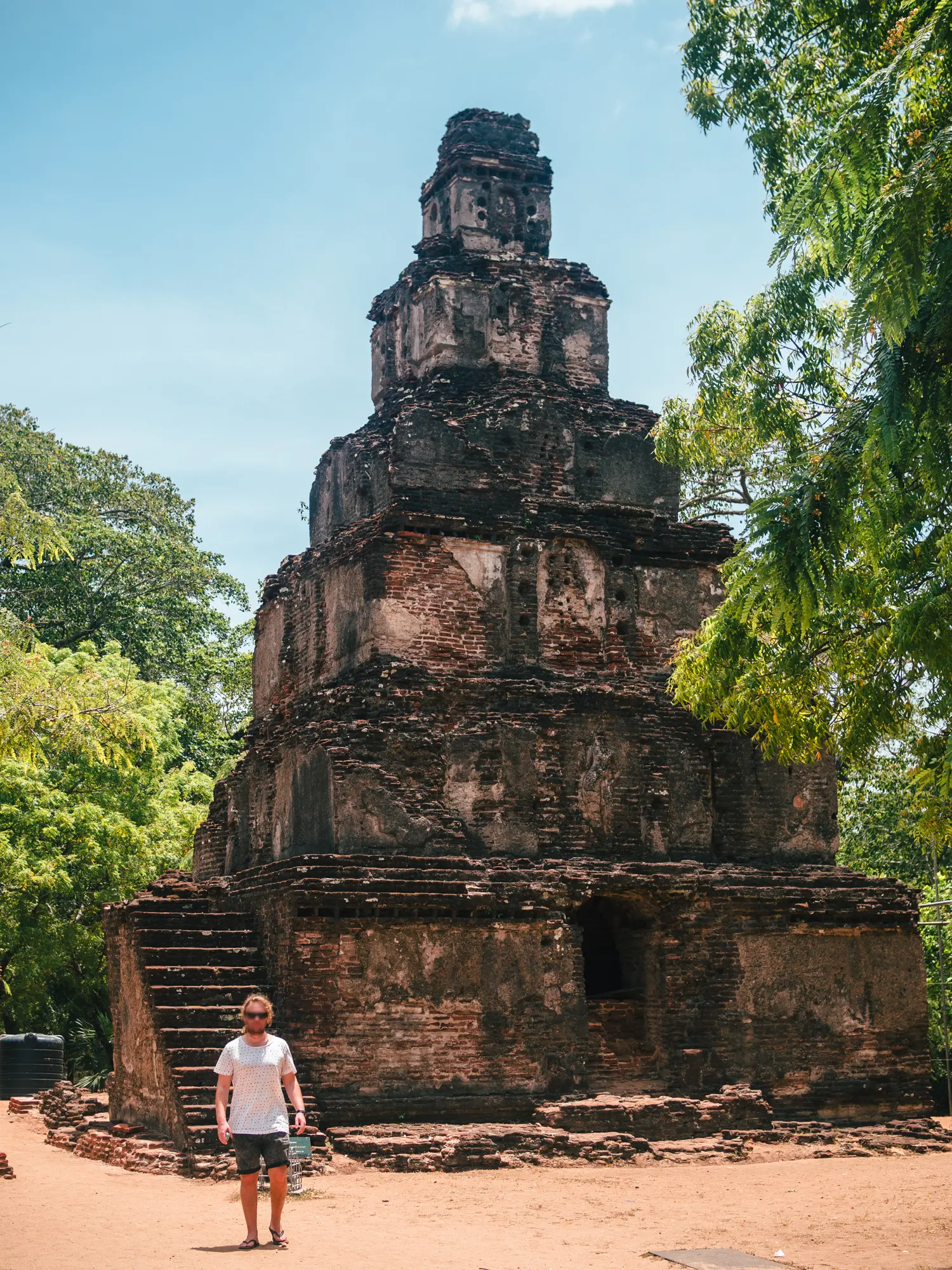 Man wearing a white t-shirt walking in front of a brick pyramid ruin with six layers, in Polonnaruwa.