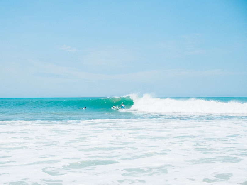 Surfer standing up on a turquoise wave with two other surfers paddling out of the way. Surfing is definitely one of the best things to do in Arugam Bay, Sri Lanka-