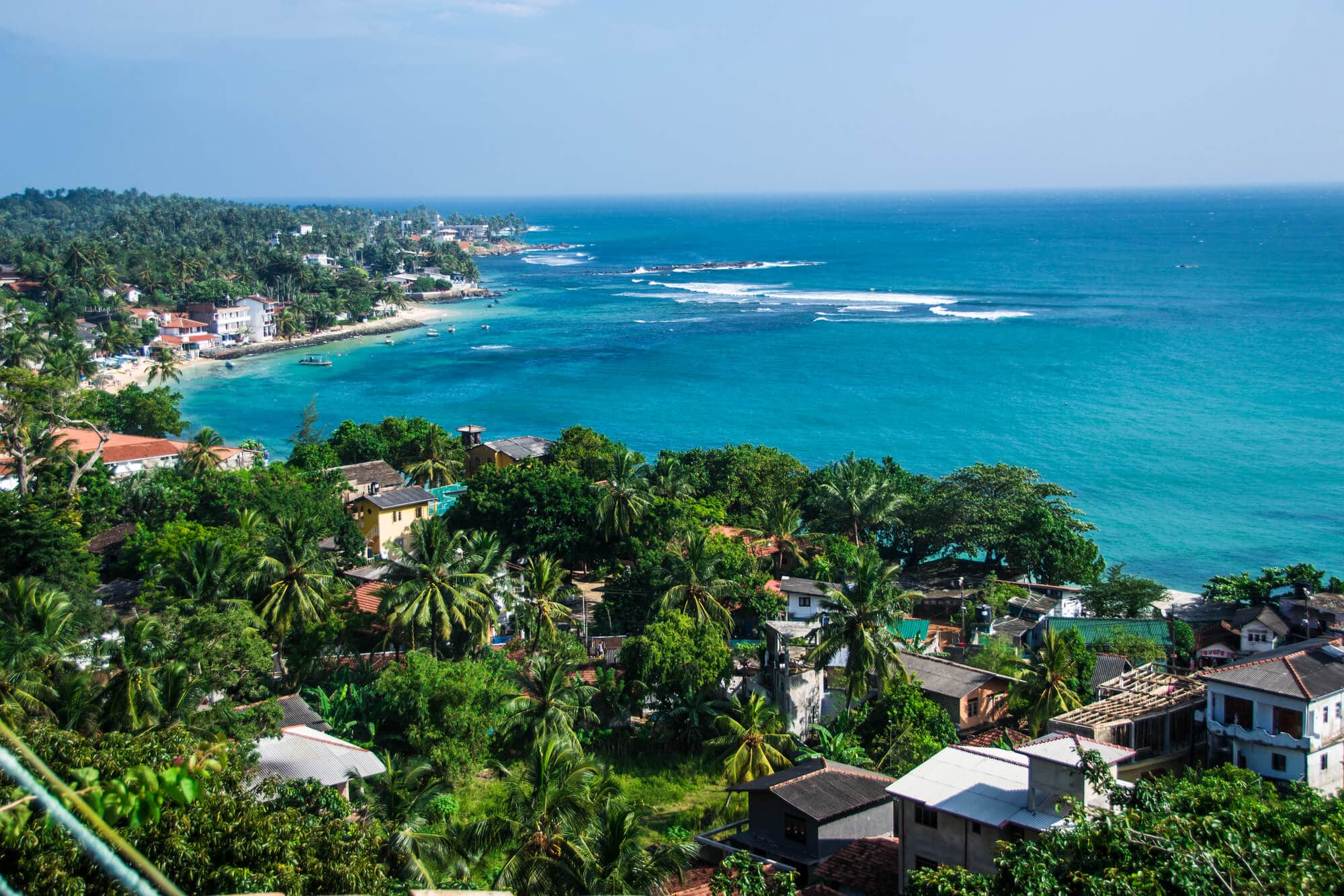 View of houses surrounded by palm trees and greenery set by the turquoise ocean from an gorgeous and affordable Airbnb in Sri Lanka.