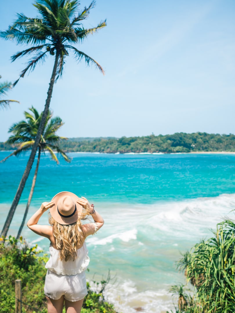 Girl with long curly hair wearing a white playsuit and beige hat looking out over the turquoise Talalla Beach Bay in Sri Lanka.