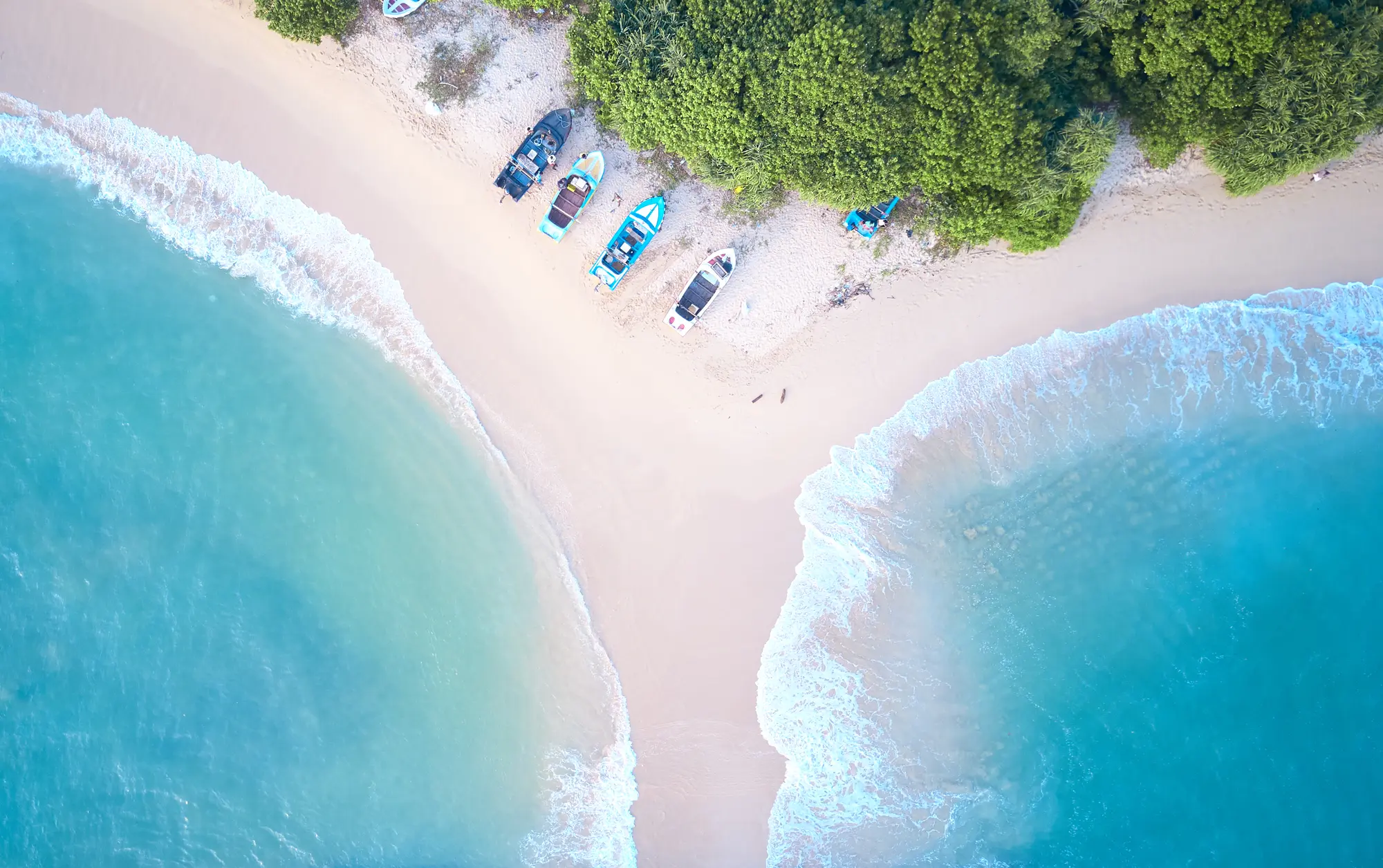 White sandbar with turquoise ocean on both sides, lined by greenery and turquoise traditional boats on Midigama Beach, a must during your 2-week Sri Lanka itinerary.