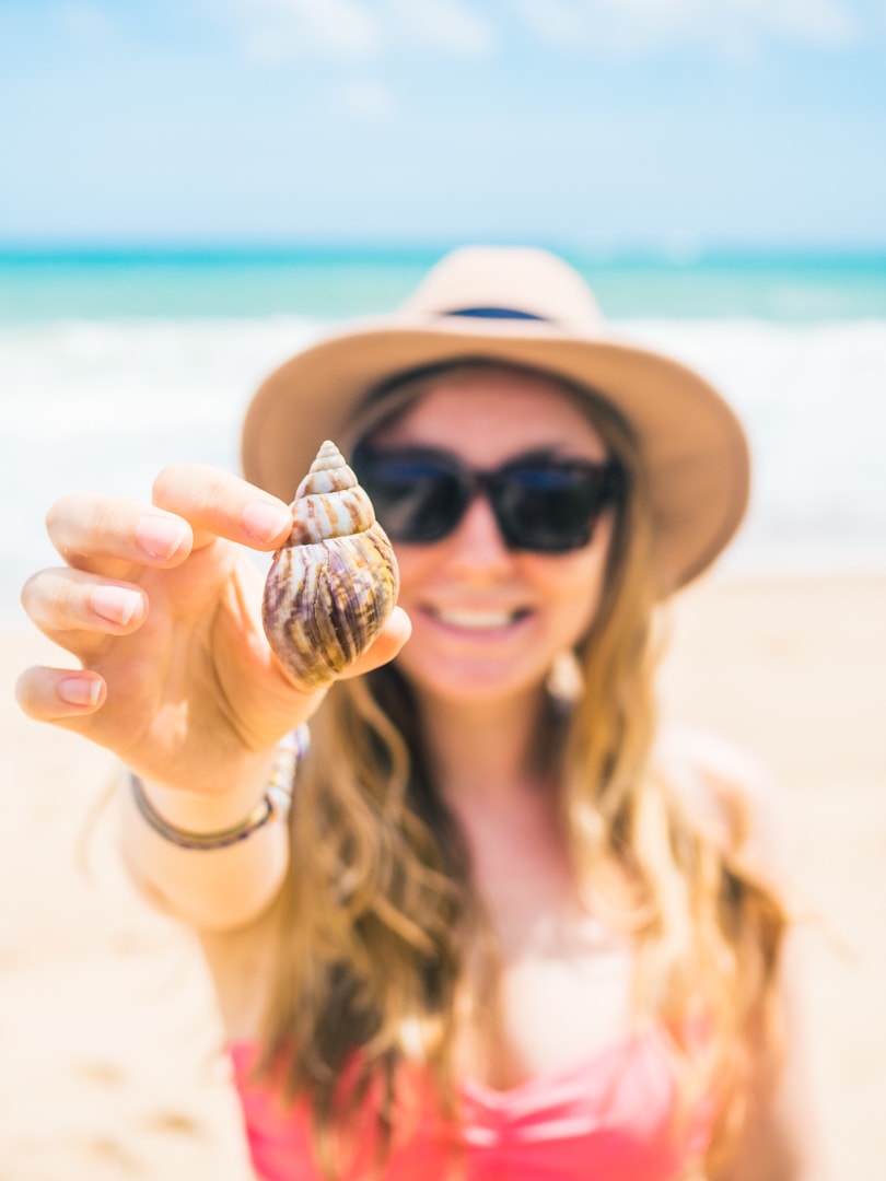 Girl wearing a hat and sunglasses holding up a white and brown seas shell on Tangalle beach, Sri Lanka.