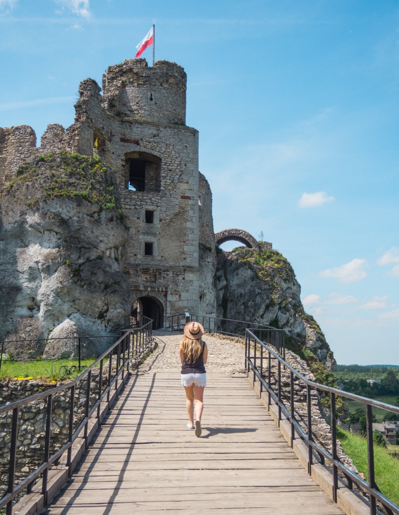 Woman with long hair, wearing a beige hat, white shorts and dark top walking on a wooden bridge towards Ogrodzieniec Castle on a sunny day during a weekend in Krakow.