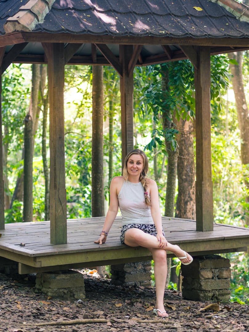 Girl sitting on a wooden gazebo, looking towards the camera, with the lush jungle in the background glowing from the afternoon light in Ubud Sacred Monkey Forest. One of the best places to see monkeys in Bali.