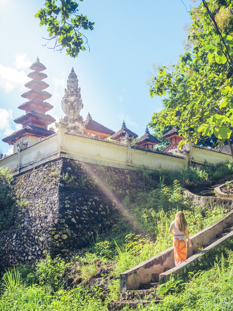 Girl wearing a white and orange sarong walking up stairs in front of traditional temple in Nusa Penida with the sun shining in.