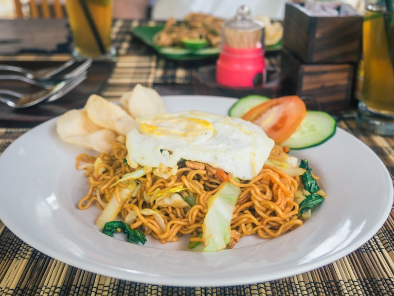 White plate with Mie Goreng fried noodles topped with an egg, cucumber and prawn crackers - Indonesian vs Thai food.