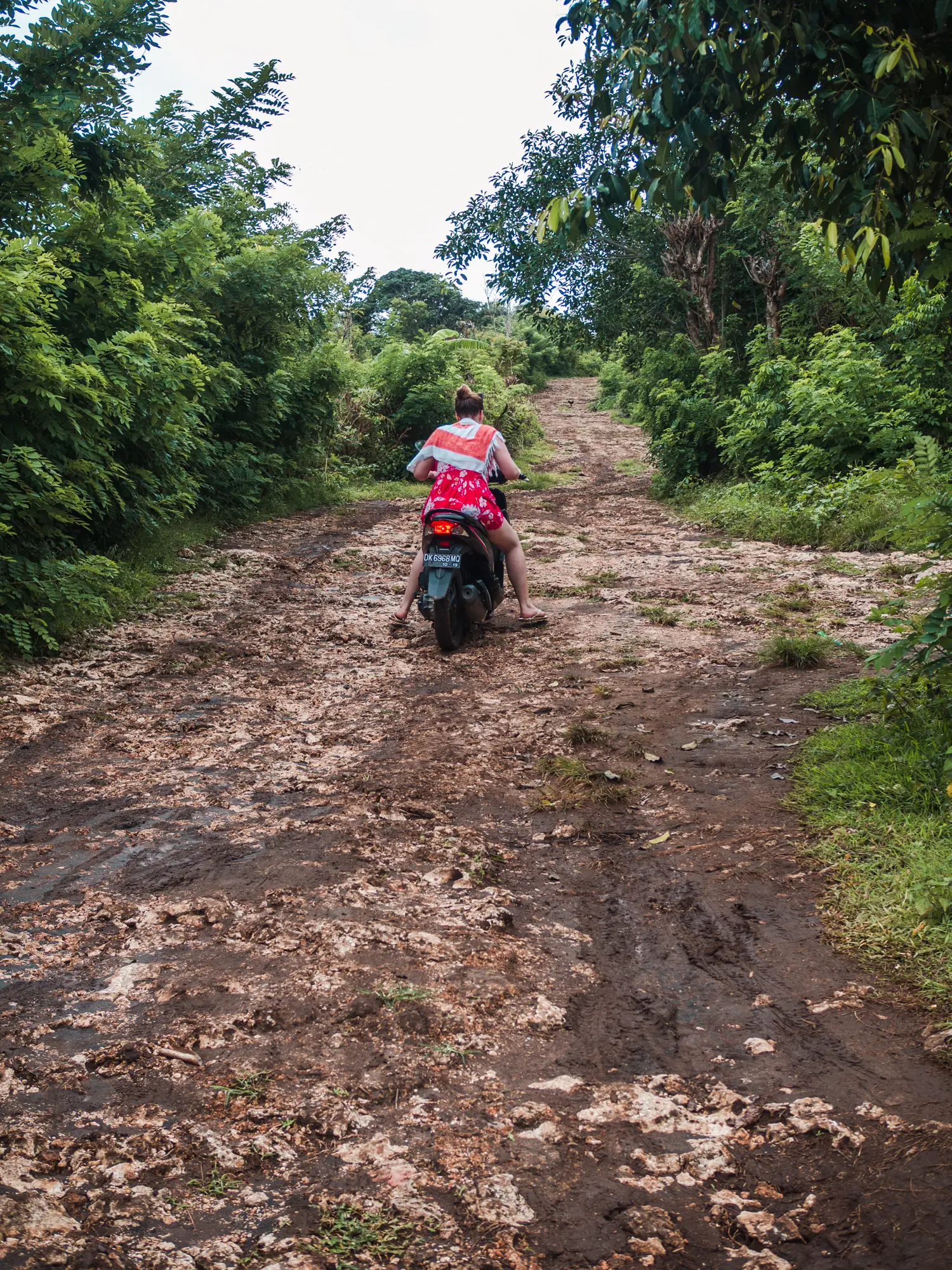 Girl in a red dress driving a scooter up s steep mud and stone road in the jungle on Nusa Penida, is it worth it?