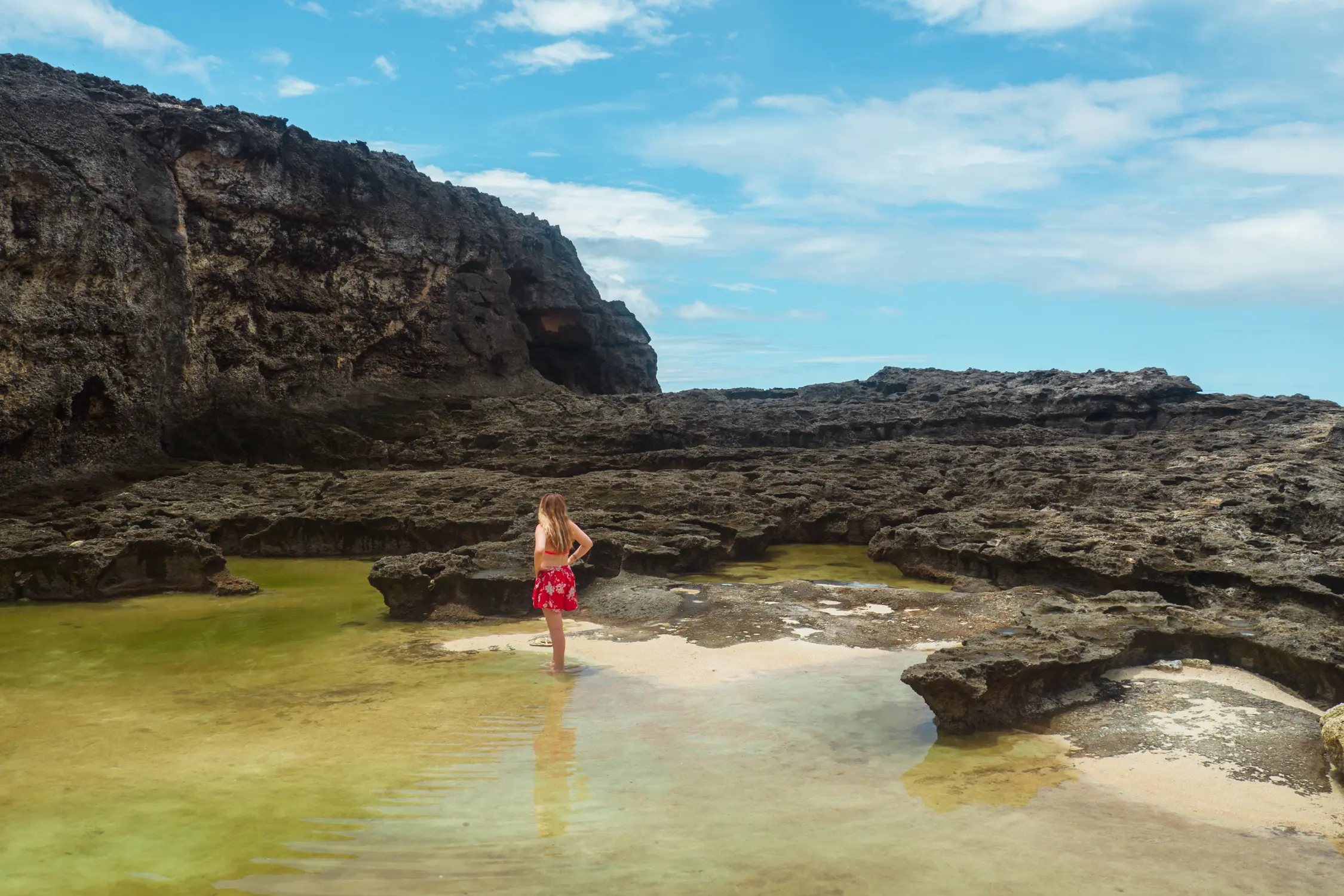 Girl in a short red dress walking in a green pond in front of volcanic rock close to Angel's Billabong, one of the main reasons to visit Nusa Penida.