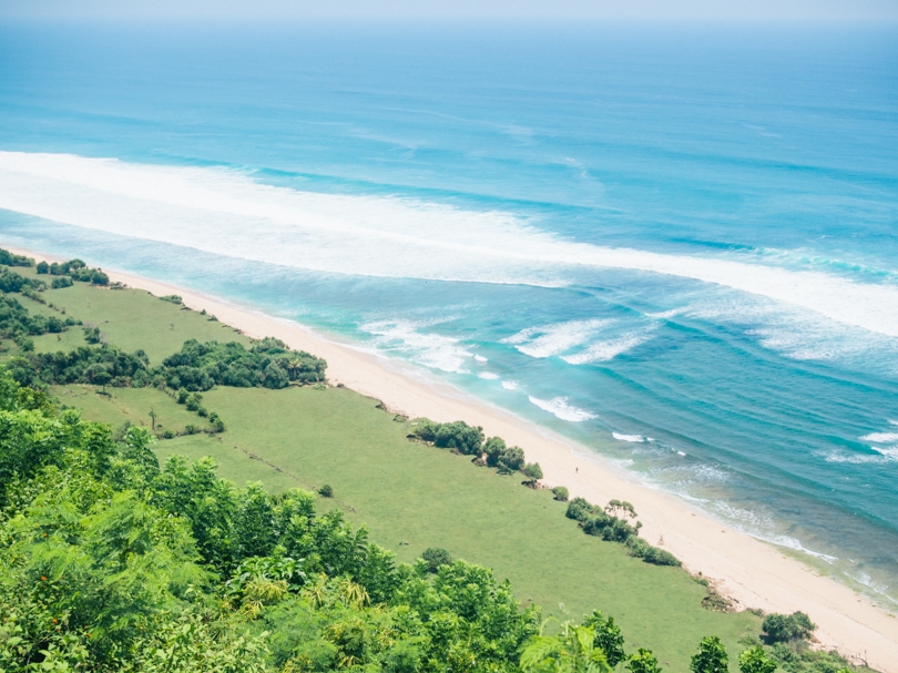 View from the tall cliff at Nyang Nyang Beach of a narrow light sand beach and green fields, one of the best beaches in Uluwatu Bali