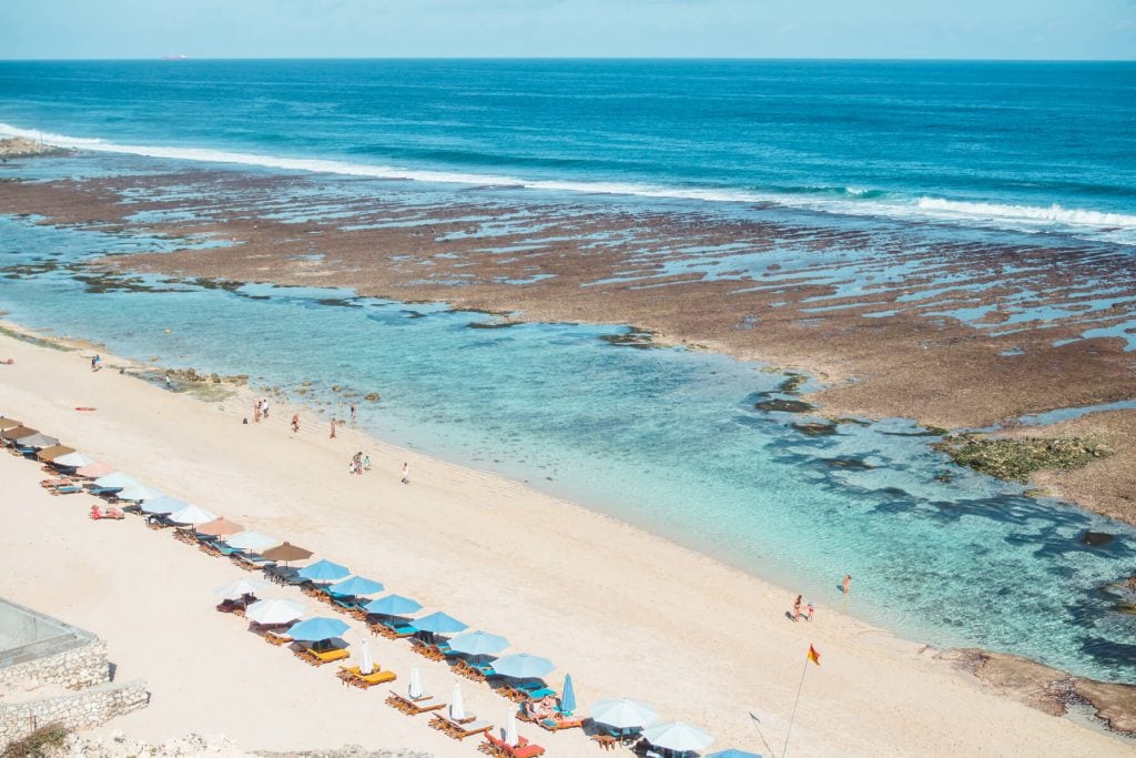 View from above of a row of umbrellas, white sand and turquoise water of Melasti Beach, on of the best beaches in Uluwatu, Bali. And a must on any two-week Bali itinerary.
