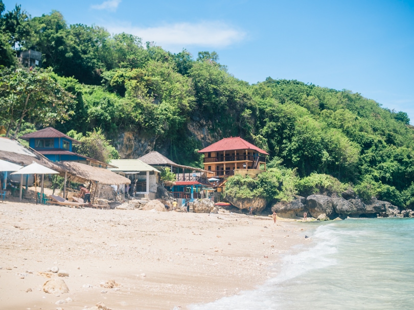 View from the water of the light sand at Thomas Beach with a view warungs and hotels set at the base of a lush cliff, one of the best beaches in Uluwatu.