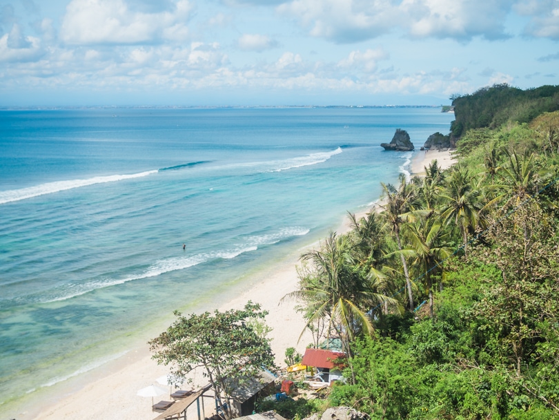 View from the cliff above of the white Thomas Beach and blue water lined by jungle and palm trees, one of the best beaches in Uluwatu Bali.