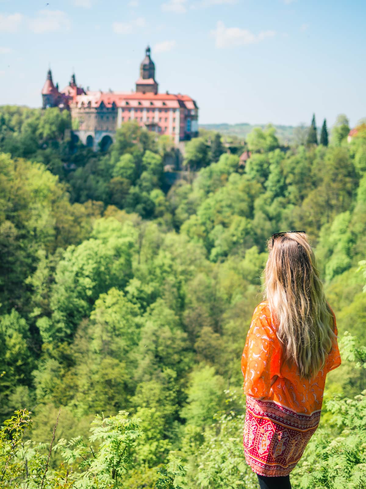 Girl in an orange kimono standing looking over at the pink Książ Castle, one of the most beautiful castles in Poland.